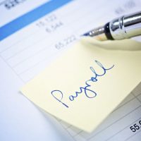 A  fountain pen checks a financial or business document with an adhesive note reading "payroll" attached to it.